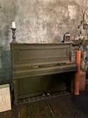 An old upright piano stands in a dimly lit room, its exterior painted in a muted green. A vintage candlestick with a white candle rests on top of the piano, alongside a lantern. A long, slender object resembling a walking cane or sword is placed across the keyboard lid. The wall behind the piano is textured, adding to the antique atmosphere. A tall orange cylinder and some decorative branches are placed to the right, while a white square panel with a simplistic key design leans against the side.