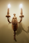 A decorative wall sconce featuring a detailed bronze sculpture of a woman with ornate leaf designs. The sconce holds two candle-shaped light bulbs that are lit, casting a warm glow and creating shadows on the wall.