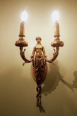 A decorative wall sconce featuring a detailed bronze sculpture of a woman with ornate leaf designs. The sconce holds two candle-shaped light bulbs that are lit, casting a warm glow and creating shadows on the wall.