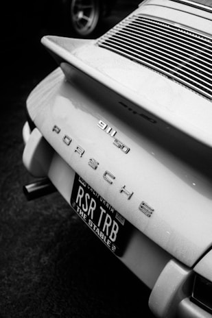 The image features the rear section of a classic Porsche 911. It displays the car's model badge '911 50' along with 'Porsche' lettering. The license plate is prominently visible with 'RSR TRB' and 'The Stable' printed on it. The scene is captured in black and white, highlighting the sleek lines and design of the vehicle.