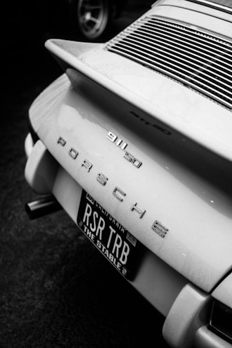 The image features the rear section of a classic Porsche 911. It displays the car's model badge '911 50' along with 'Porsche' lettering. The license plate is prominently visible with 'RSR TRB' and 'The Stable' printed on it. The scene is captured in black and white, highlighting the sleek lines and design of the vehicle.