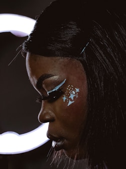A close-up of a person in profile, showcasing elaborate makeup with artistic designs of stars and lines on their face, highlighted by a dramatic contrast of light and shadow. The person has dark hair, styled neatly, with a hairpin visible.