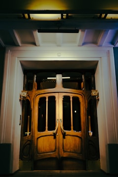 An ornate wooden door with intricate glass panels set in a well-lit entrance. The framing of the doorway is structured and elegant, with warm lighting highlighting the details of the woodwork.