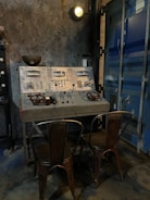 A vintage industrial control panel with numerous dials, switches, and gauges is mounted on a rough, concrete-style wall. Two distressed metal chairs are placed in front of the panel. The scene is dimly lit by an overhead industrial light. Adjacent to the control panel is the metallic texture of a blue industrial door.