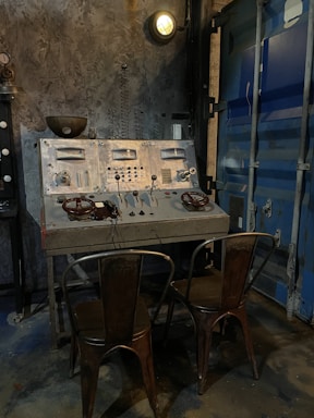 A vintage industrial control panel with numerous dials, switches, and gauges is mounted on a rough, concrete-style wall. Two distressed metal chairs are placed in front of the panel. The scene is dimly lit by an overhead industrial light. Adjacent to the control panel is the metallic texture of a blue industrial door.