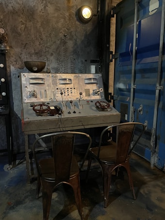 A vintage industrial control panel with numerous dials, switches, and gauges is mounted on a rough, concrete-style wall. Two distressed metal chairs are placed in front of the panel. The scene is dimly lit by an overhead industrial light. Adjacent to the control panel is the metallic texture of a blue industrial door.