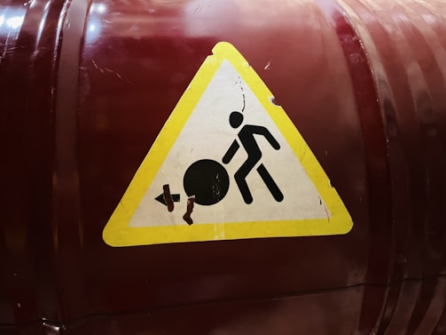 A caution sign with a yellow triangular background featuring a black stick figure pushing a large black circle. The circle has a crack, indicating a potential hazard. The sign is affixed to a dark red and slightly reflective cylindrical surface.
