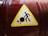 A caution sign with a yellow triangular background featuring a black stick figure pushing a large black circle. The circle has a crack, indicating a potential hazard. The sign is affixed to a dark red and slightly reflective cylindrical surface.