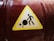 A caution sign with a yellow triangular background featuring a black stick figure pushing a large black circle. The circle has a crack, indicating a potential hazard. The sign is affixed to a dark red and slightly reflective cylindrical surface.