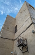 A large, beige, textured building wall with visible wires running horizontally across it. An air conditioning unit is mounted within a metal cage, secured to the wall. The image includes a single wall-mounted light fixture and features a clear blue sky with scattered clouds in the background.