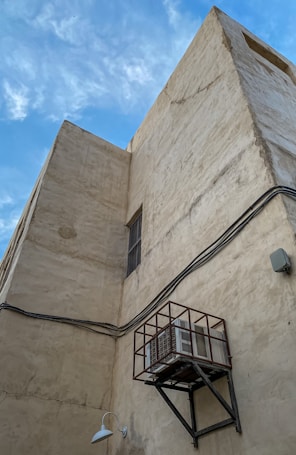 A large, beige, textured building wall with visible wires running horizontally across it. An air conditioning unit is mounted within a metal cage, secured to the wall. The image includes a single wall-mounted light fixture and features a clear blue sky with scattered clouds in the background.