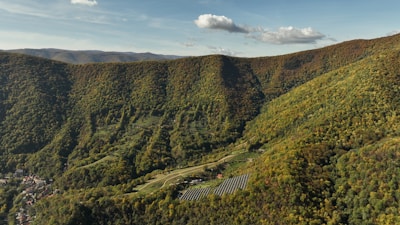 Aerial view of a lush, forested mountain landscape with a small village at the foot of the hills. Among the green foliage, a series of solar panels are visible, adding a modern touch to the natural setting. The sky above is clear with a few scattered clouds, enhancing the serene atmosphere.