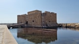 A historic stone fortress stands beside a calm body of water, connected by a small stone bridge with a wooden railing. The fortress, constructed from large, aged bricks, shows the wear of time, giving it a rustic appearance. The surrounding water is clear, reflecting parts of the fortress and the sky. In the background, a clear blue sky complements the tranquil scene.