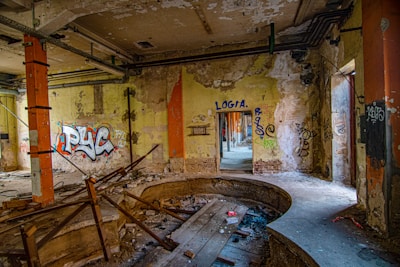 An abandoned and dilapidated interior with graffiti on the walls, exposed pipes, and visible structural damage. Debris and remnants of construction materials are scattered across the floor, and the room has an overall neglected and decayed appearance.