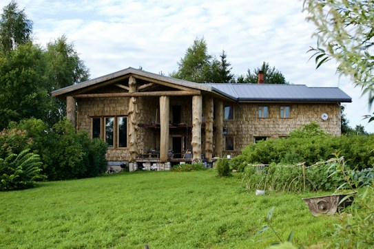 A rustic log cabin with a modern roof is surrounded by lush greenery and a neatly mowed lawn. The facade features large windows and distinctive wooden pillars. The setting is serene, with trees and shrubs enhancing the natural landscape.