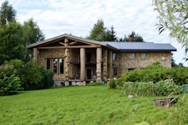 A rustic log cabin with a modern roof is surrounded by lush greenery and a neatly mowed lawn. The facade features large windows and distinctive wooden pillars. The setting is serene, with trees and shrubs enhancing the natural landscape.