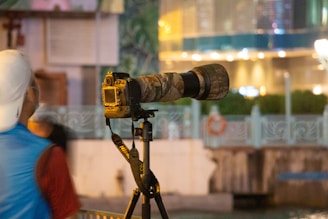 A professional camera mounted on a tripod with a large telephoto lens is positioned for photography. The setting appears to be outdoors with decorative elements like a patterned railing and foliage visible in the background. A person wearing a cap and a blue vest stands nearby.
