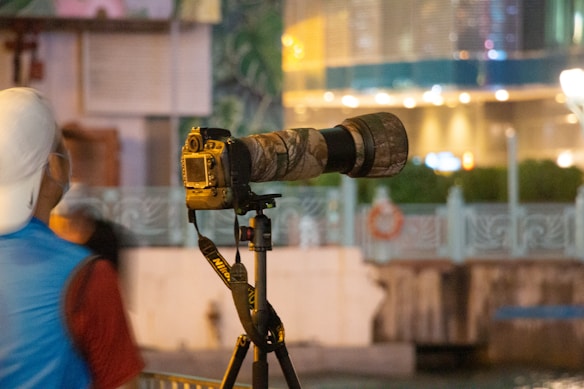 A professional camera mounted on a tripod with a large telephoto lens is positioned for photography. The setting appears to be outdoors with decorative elements like a patterned railing and foliage visible in the background. A person wearing a cap and a blue vest stands nearby.