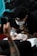 A person is receiving a tattoo on their arm by a tattoo artist wearing gloves and a face mask. The tattoo artist is focused on the task, using a tattoo machine. The background is dark, with some blue and turquoise elements.
