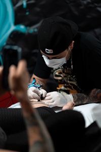 A person is receiving a tattoo on their arm by a tattoo artist wearing gloves and a face mask. The tattoo artist is focused on the task, using a tattoo machine. The background is dark, with some blue and turquoise elements.