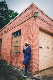 A person dressed in a blue tracksuit with red stripes leans casually against the corner of a brick building. They wear a head covering and sandals, and appear relaxed. The building features old windows, a garage door, and a weathered exterior. The surrounding area shows green foliage and a concrete ground.