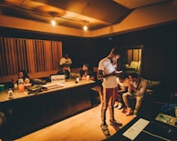 A recording studio scene with several people interacting. One person is standing and looking at a smartphone, while others are seated at a mixing console or on couches. The room is warmly lit with wood paneling and studio equipment visible.