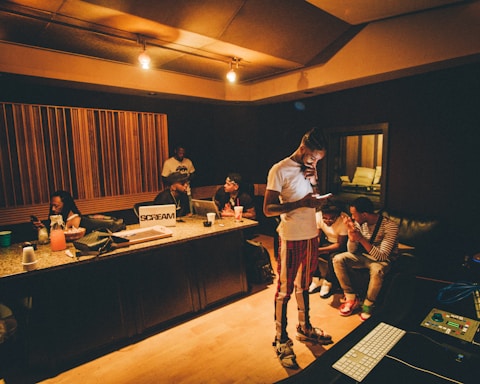 A recording studio scene with several people interacting. One person is standing and looking at a smartphone, while others are seated at a mixing console or on couches. The room is warmly lit with wood paneling and studio equipment visible.