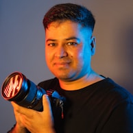 A person is holding a professional camera with a lens that has a red glow. The lighting creates a dramatic contrast on their face with warm and cool tones.