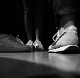 Several pairs of sneakers are visible at a low angle, with the focus on a close-up of a Nike shoe on the right side. The background shows other shoes and the legs of individuals, all depicted in black and white.