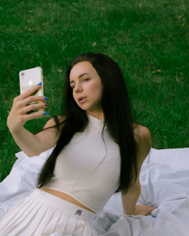A young dark-haired woman reclining on a white cloth on grass, holding a clear-cased iPhone up in front of her and looking into its camera to take