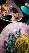 A delectable seafood pasta dish is served in a rustic ceramic bowl with a side of mussels and calamari. The spaghetti is accompanied by small, colorful clams, adding vibrancy to the meal. The table also features additional ceramic bowls in a teal hue, enhancing the overall aesthetic.