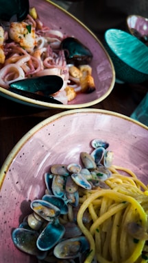 A delectable seafood pasta dish is served in a rustic ceramic bowl with a side of mussels and calamari. The spaghetti is accompanied by small, colorful clams, adding vibrancy to the meal. The table also features additional ceramic bowls in a teal hue, enhancing the overall aesthetic.
