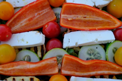 A vibrant assortment of fresh salad ingredients including red and yellow cherry tomatoes, sliced cucumbers, pieces of feta cheese, and halved red bell peppers. The elements are arranged closely together, showcasing their natural colors and textures.