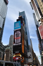 Tall skyscrapers and numerous electronic billboards featuring various advertisements dominate an urban scene. A prominent advertisement for a beverage and a Dunkin' ad stand out. Busy streets with vehicles are visible at the bottom of the image.