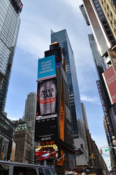 Tall skyscrapers and numerous electronic billboards featuring various advertisements dominate an urban scene. A prominent advertisement for a beverage and a Dunkin' ad stand out. Busy streets with vehicles are visible at the bottom of the image.