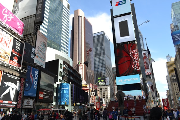 A bustling urban scene filled with tall skyscrapers and numerous billboards advertising various brands such as Coca-Cola, Hyundai, and a musical show among others. People are walking around in the area, and the scene includes a red staircase leading upward, adding a focal point. The setting is busy with bright lights and advertisements dominating the view.