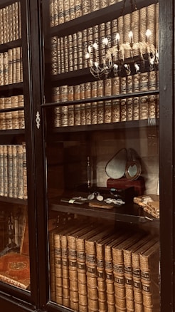 A glass-fronted bookcase filled with vintage leather-bound books on wooden shelves, adorned with ornate gold patterns. A decorative chandelier with lit candles is reflected in the glass. There is also a small selection of vintage items on display, including a pair of spectacles and a magnifying glass, resting on a shelf along with a framed oval mirror.
