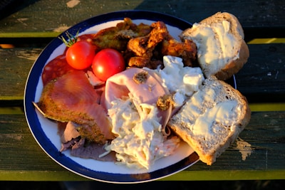 A plate featuring a variety of food items including slices of smoked salmon, roast beef, ham, coleslaw, cherry tomatoes, bread with butter, and spiced chicken pieces. The plate is set on a wooden table, suggesting an outdoor setting.