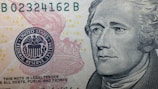 A close-up of a US Federal Reserve Note featuring intricate line work and detailed imagery. The portrait of a historical figure is prominent on the right, while a portion of the Federal Reserve emblem with an eagle is visible on the left. Small '10' numerals are scattered across the background.