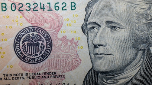 A close-up of a US Federal Reserve Note featuring intricate line work and detailed imagery. The portrait of a historical figure is prominent on the right, while a portion of the Federal Reserve emblem with an eagle is visible on the left. Small '10' numerals are scattered across the background.