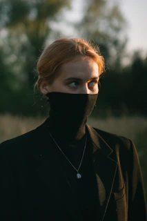 A person with light brown hair is wearing a dark turtleneck sweater that covers their mouth and nose, along with a dark blazer. They are standing outdoors with a blurred background of green foliage. The lighting is soft and warm, suggesting it is either early morning or late afternoon.