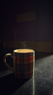 A ceramic mug with a plaid pattern in red, blue, and black sits on a dark, speckled surface. Soft light creates a warm glow around the mug, highlighting steam rising from the hot beverage inside.