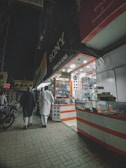 A nighttime street scene with two people walking past an electronics shop displaying various gadgets and accessories. Bright lights illuminate the shop interior, with signage visible. The sidewalk is paved, and a motorcycle is parked nearby.