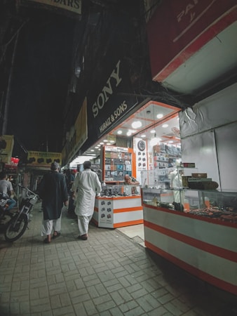 A nighttime street scene with two people walking past an electronics shop displaying various gadgets and accessories. Bright lights illuminate the shop interior, with signage visible. The sidewalk is paved, and a motorcycle is parked nearby.