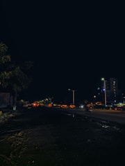 A nighttime urban scene featuring a street with moving traffic, streetlights, and adjacent buildings. The foreground shows a dark road with visible tire tracks, while the background displays illuminated vehicles and high-rise structures.