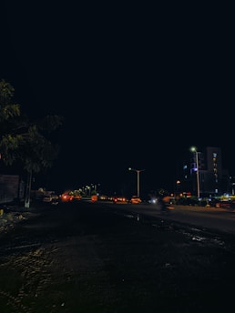 A nighttime urban scene featuring a street with moving traffic, streetlights, and adjacent buildings. The foreground shows a dark road with visible tire tracks, while the background displays illuminated vehicles and high-rise structures.