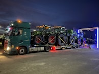 A large truck carries three tractors on its flatbed, parked in an open area during the evening. The sky is dark and the truck's lights are illuminated, casting a glow around. The tractors are green and securely strapped onto the truck.