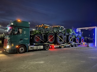 A large truck carries three tractors on its flatbed, parked in an open area during the evening. The sky is dark and the truck's lights are illuminated, casting a glow around. The tractors are green and securely strapped onto the truck.