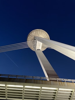 The image features a modern architectural structure with tall, steel supports converging towards a circular platform at the top. The structure appears to be part of a bridge, with cables extending outward. The background shows a clear, deep blue sky.