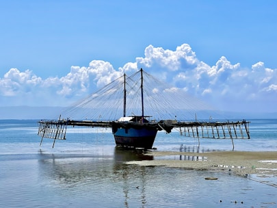 A unique fishing structure is positioned on a sandy shore, with a large, net-like apparatus extending over the water. It's under a bright blue sky with scattered fluffy clouds. The structure has a central mast and numerous cables stretching outwards over the sea.